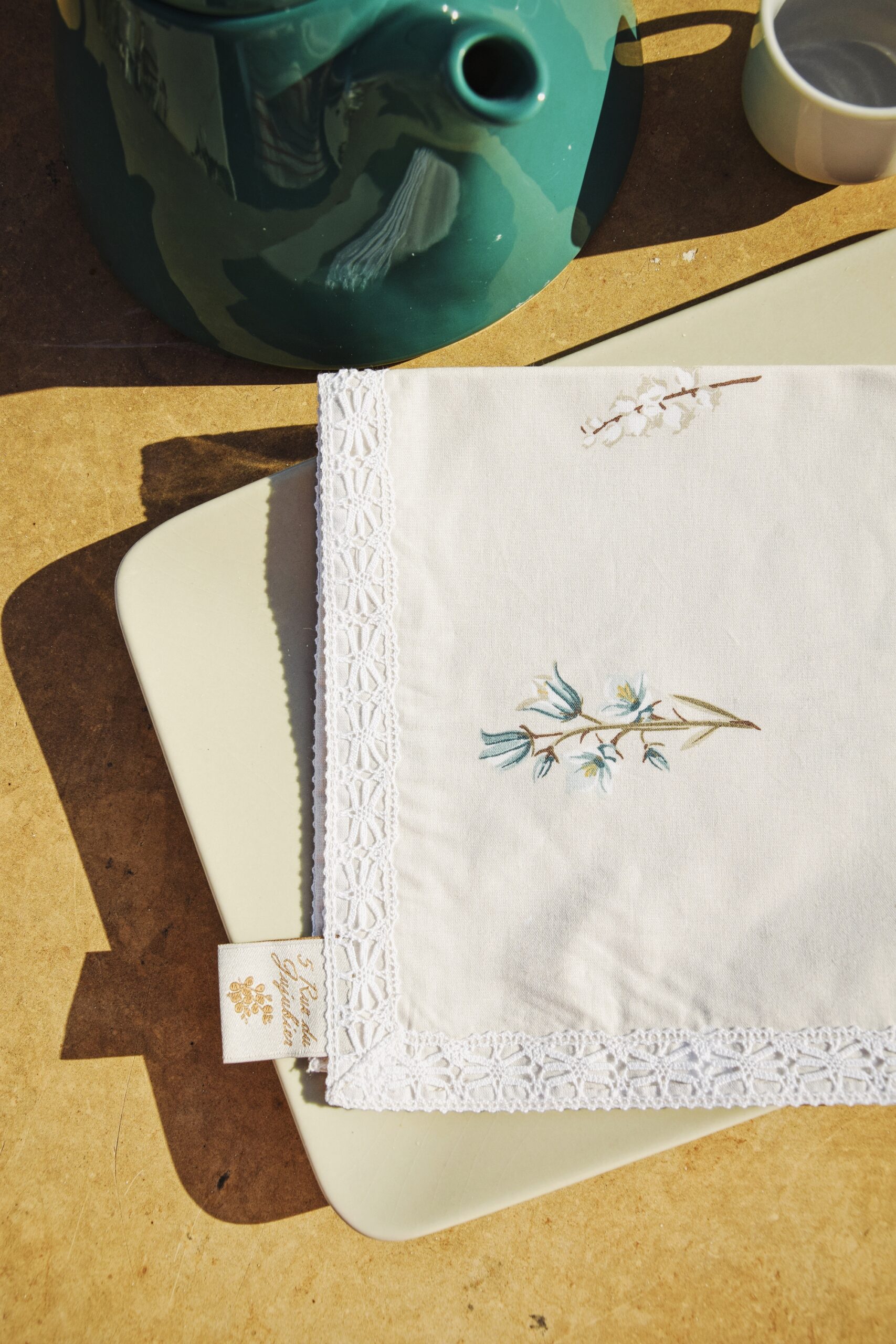 Serviette brodée dentelle Tulipe Dentelle avec motif floral bleu-vert et bordure guipure, posée sur une table en bois